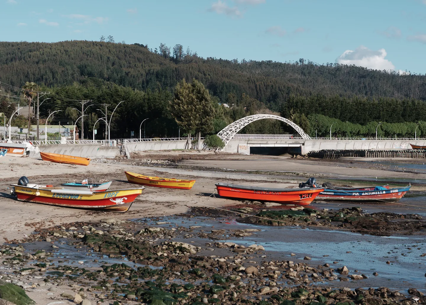 Fishing boats on the beach at low tide, with a white arch bridge and wooded hills behind.